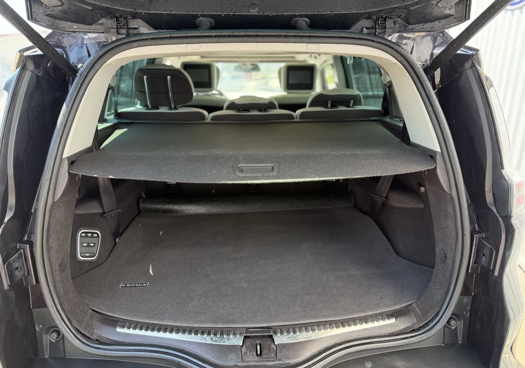 Vue arrière du coffre ouvert du Renault Espace V noir, avec cache-bagages déployé et sièges arrière équipés d'écrans intégrés.