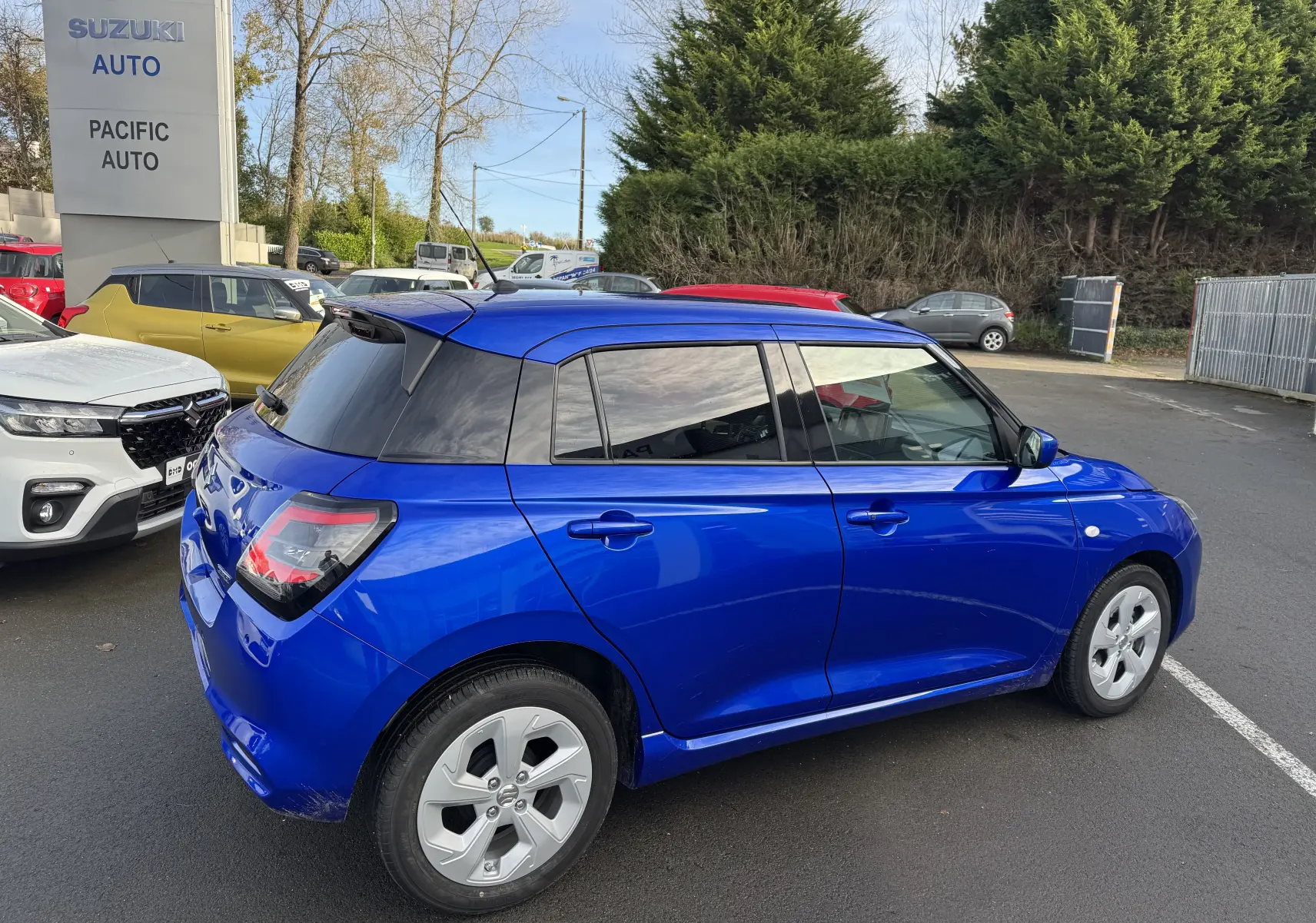 Vue 3/4 arrière droite d'une Suzuki Swift 1.2 Hybrid Privilege Auto bleu avec vitres teintées sur parking extérieur.