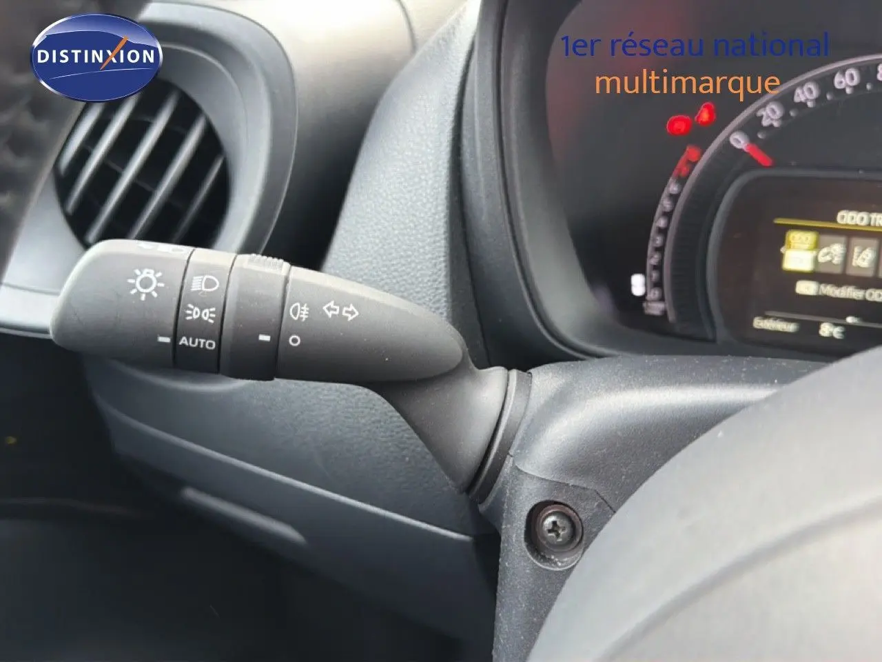 Gros plan sur la commande des phares et clignotants à gauche du volant d’une Toyota Aygo X noire, tableau de bord partiellement visible.