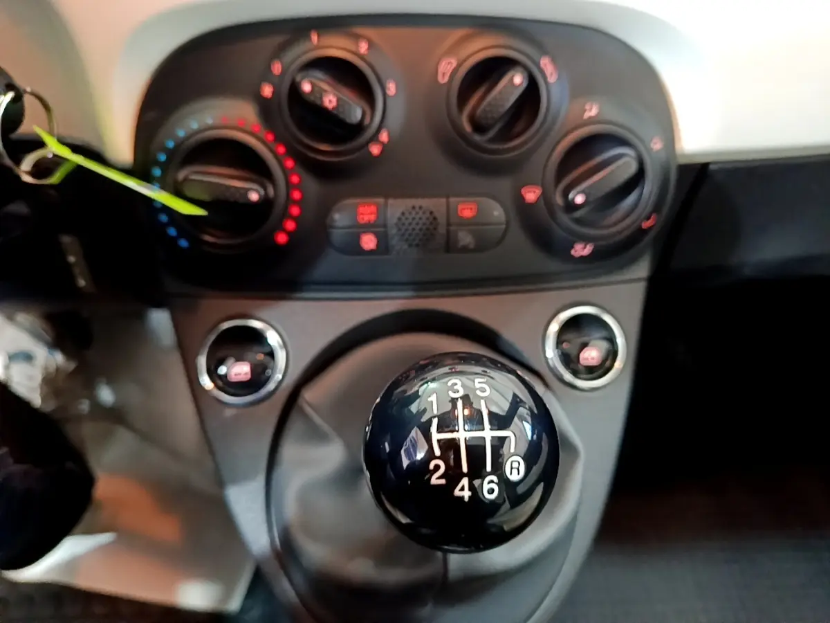 Gros plan sur le levier de vitesses manuel noir et la console centrale avec commandes de climatisation d'une Fiat 500 blanche.