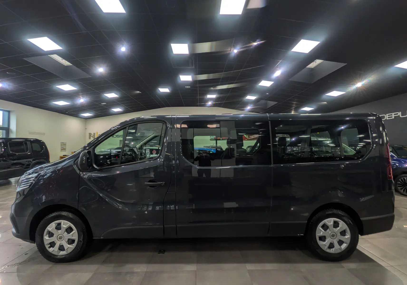 Profil droit du Renault Trafic Combi L2 gris Comète 2023 en intérieur, mettant en valeur ses grandes fenêtres et sa silhouette allongée.