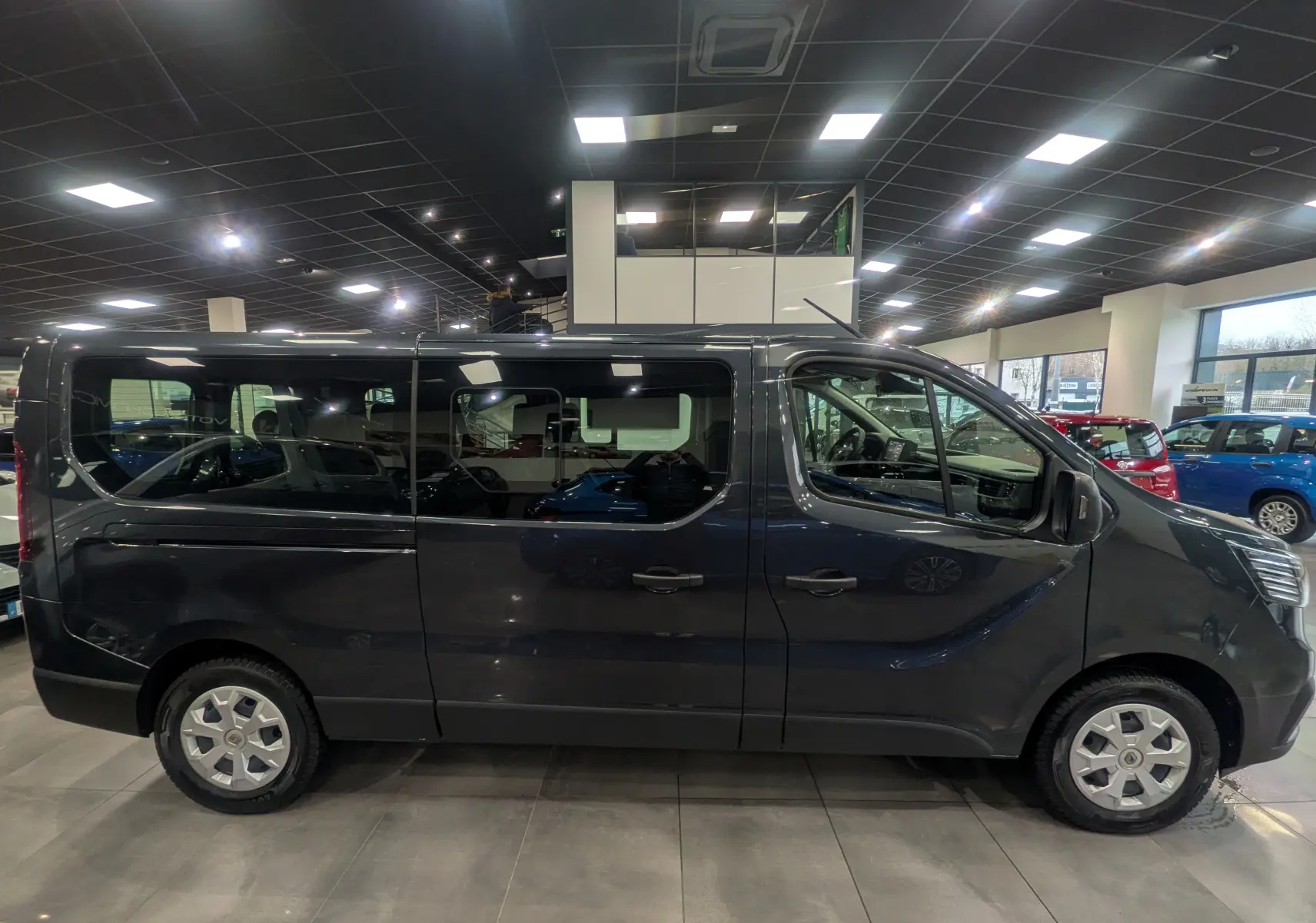 Vue de profil côté gauche du Renault Trafic Combi L2 gris comète 2023 en intérieur, avec fenêtres teintées et jantes acier.