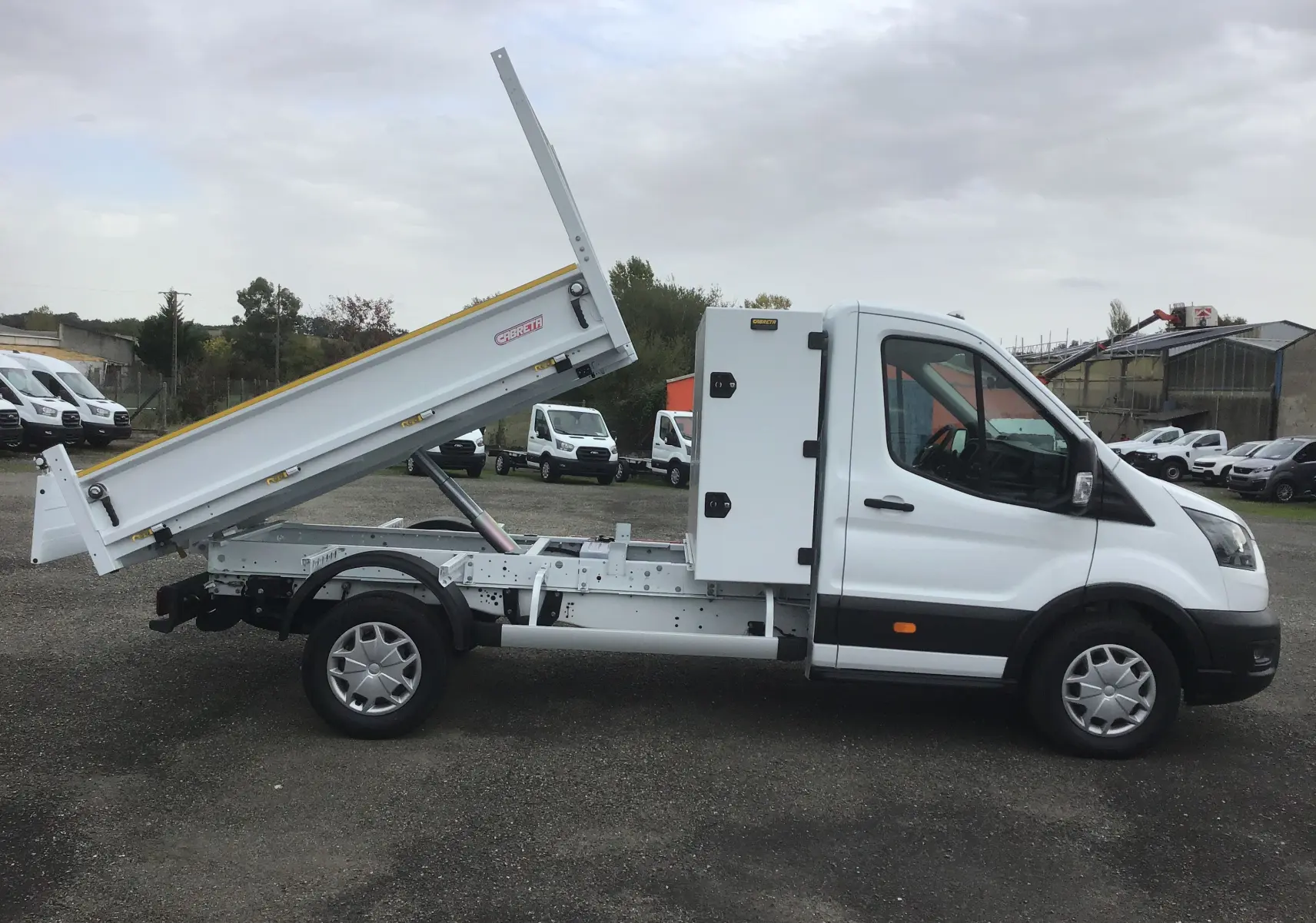 Vue de profil côté gauche d'un Ford Transit blanc avec benne relevée et coffre latéral acier.