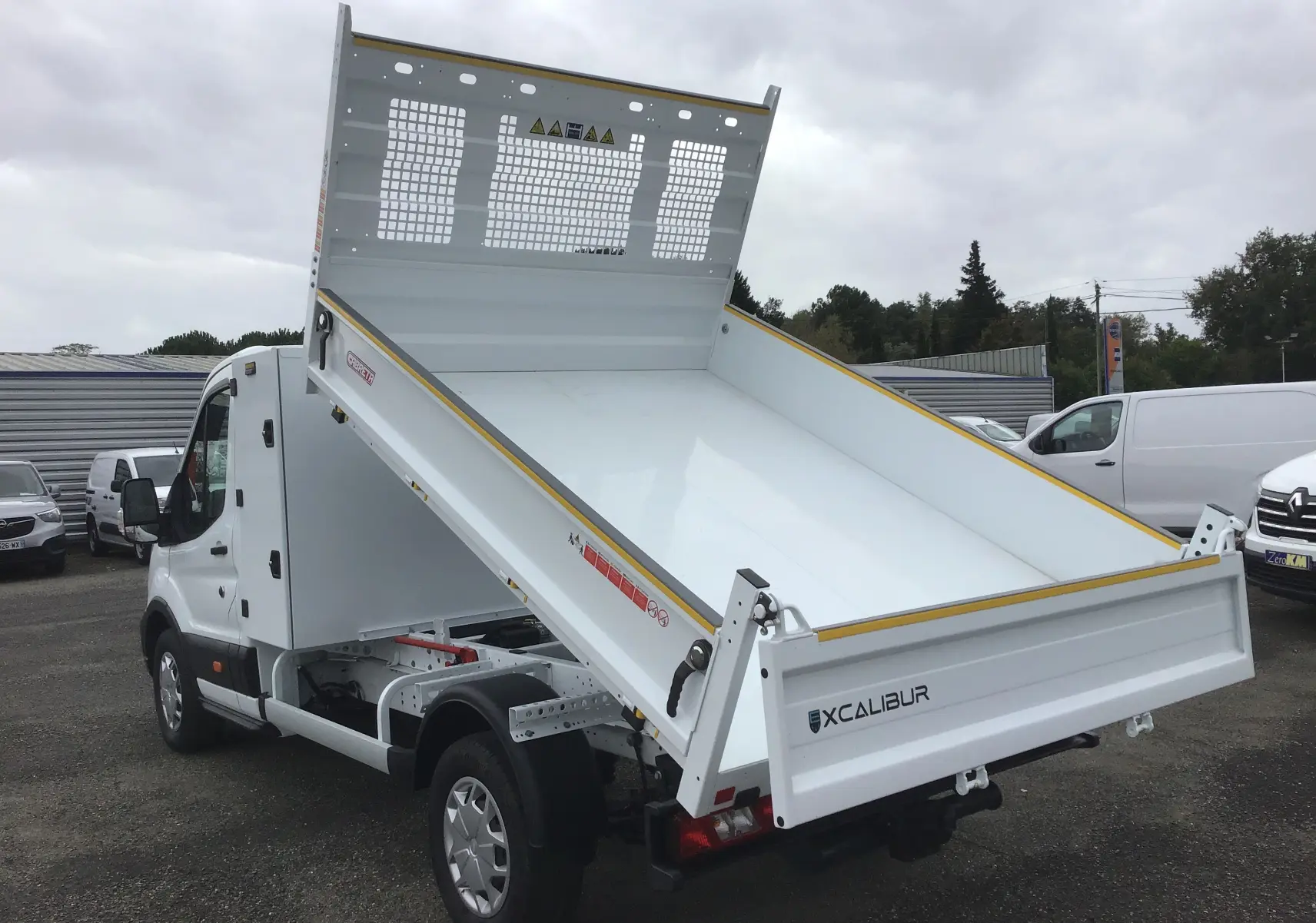Vue 3/4 arrière droite du Ford Transit Châssis Cabine blanc avec benne relevée et coffre acier visible.