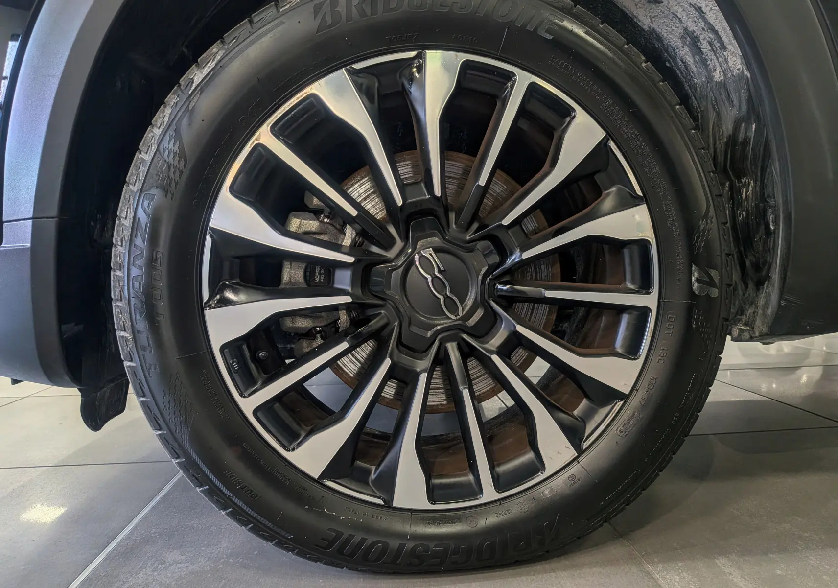 Gros plan sur la roue avant droite du FIAT 500X 2024 noir Cinéma, avec jante alliage bicolore et pneu Bridgestone.