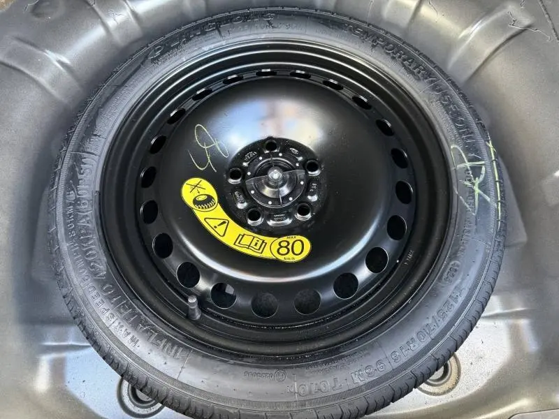Pneu de secours noir avec étiquette jaune indiquant une vitesse maximale de 80 km/h, logé dans le coffre d'une Ford Focus gris lunaire.