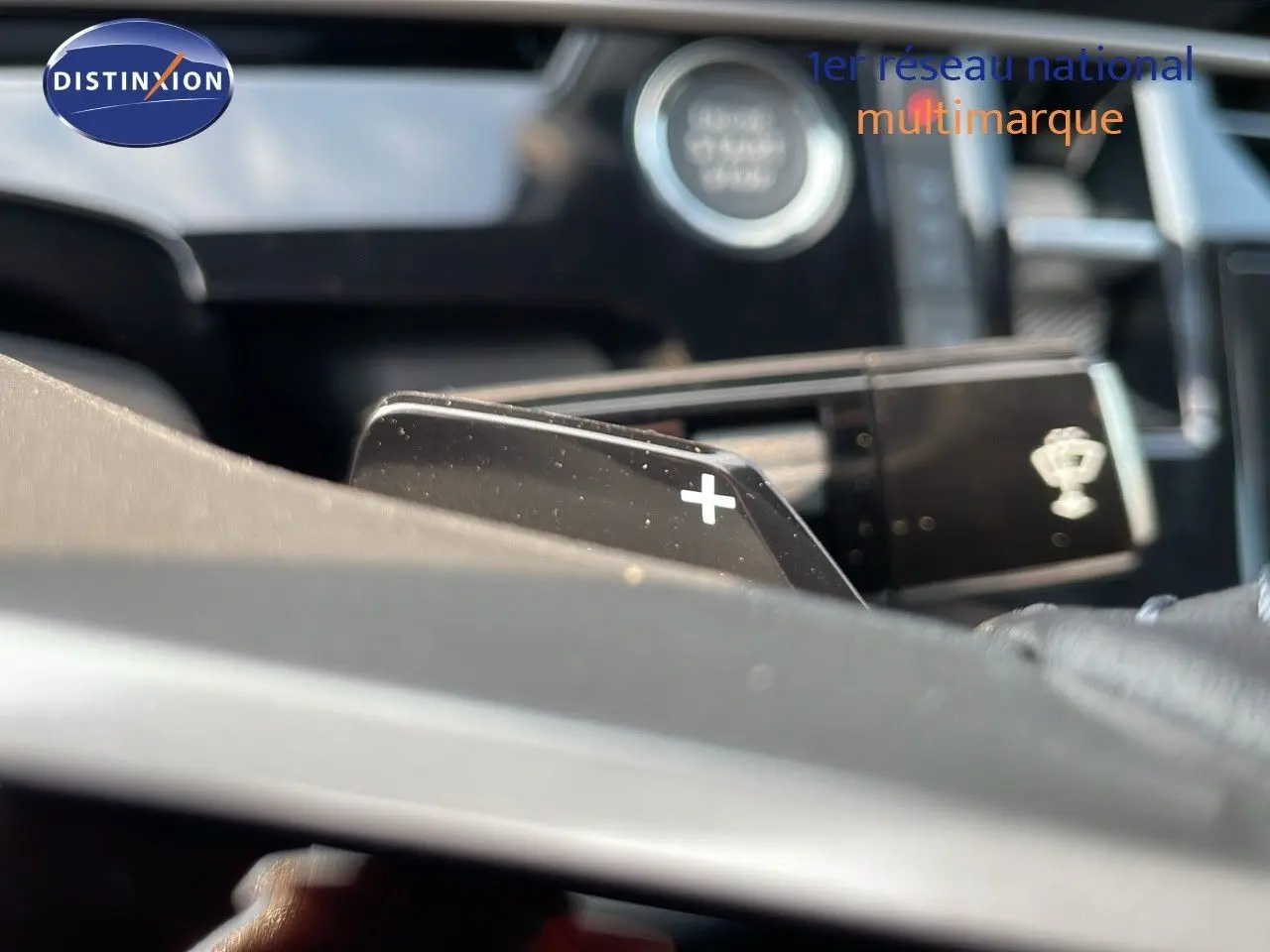 Gros plan sur la palette de changement de vitesse "+" du Peugeot 3008 gris, avec bouton start visible en arrière-plan flou.