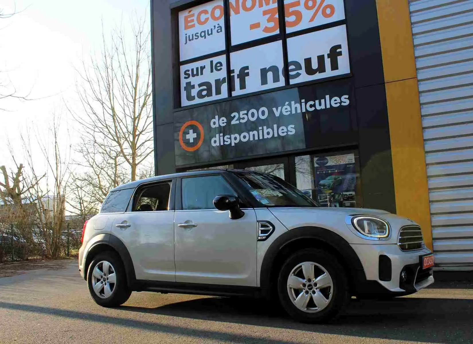 Vue latérale gauche d'un MINI Countryman gris perle métal avec toit noir, stationné devant une enseigne commerciale.