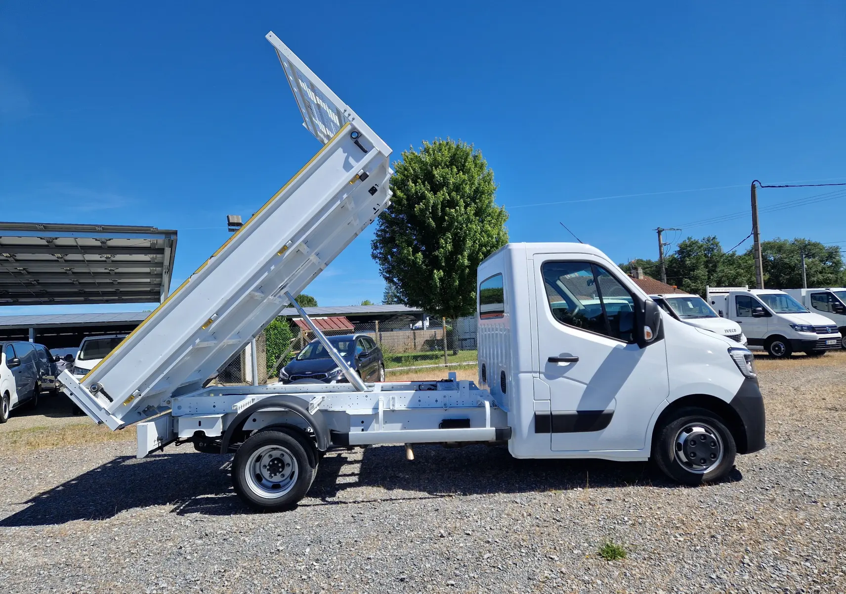 Vue de profil côté droit d’un Renault Master blanc avec benne basculante relevée sur un terrain extérieur.