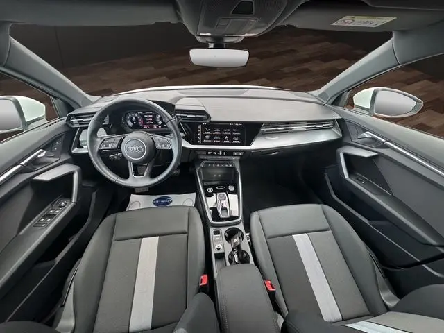 Vue intérieure avant de l'Audi A3 Sportback 2024 avec tableau de bord digital, volant cuir multifonctions et sièges tissu noir.