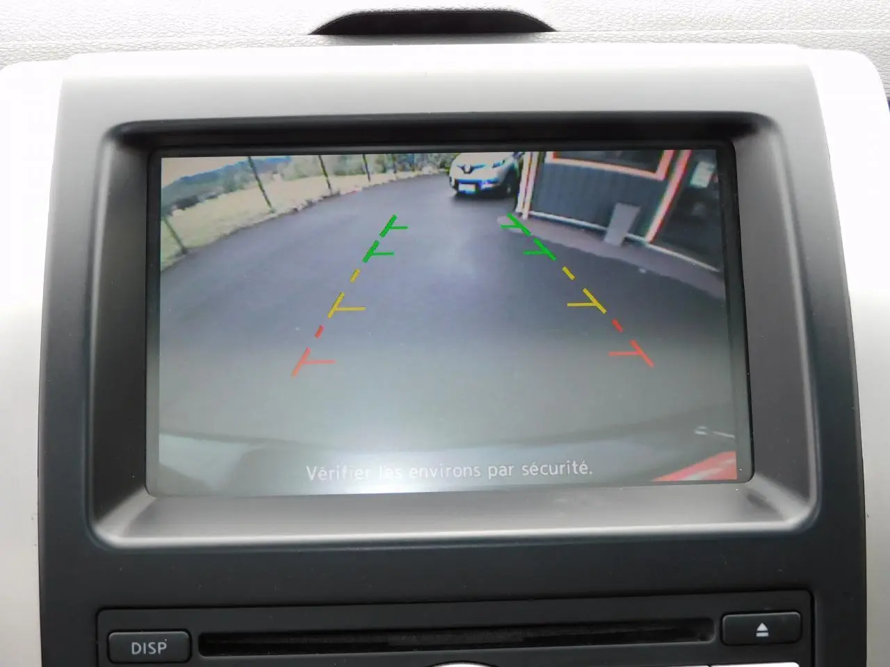 Écran de caméra de recul du Nissan X-TRAIL noir montrant l’arrière avec lignes de guidage colorées et un véhicule blanc en fond.