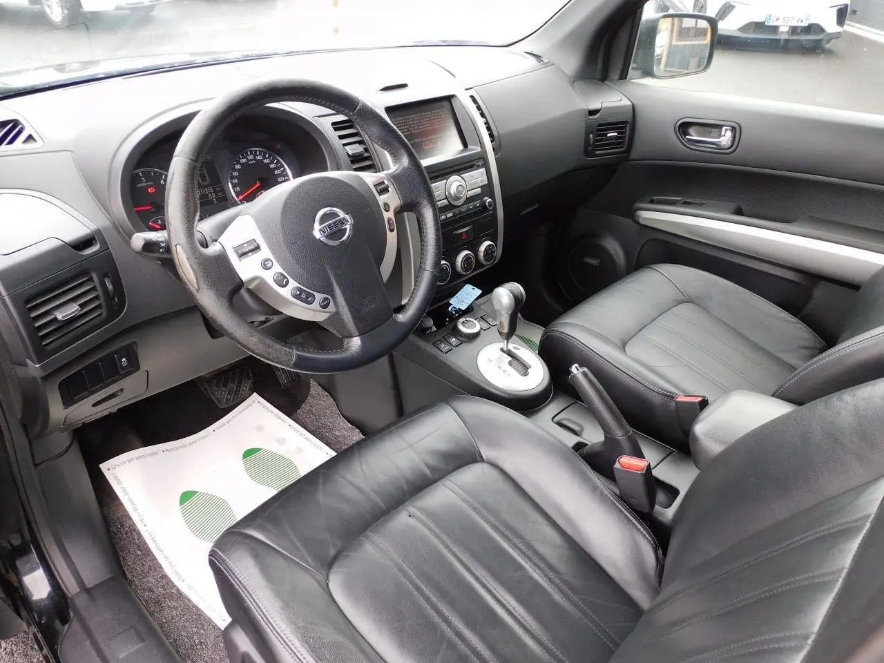 Vue intérieure du Nissan X-TRAIL 2011 montrant les sièges cuir noirs, le volant multifonction et la console centrale avec boîte auto.