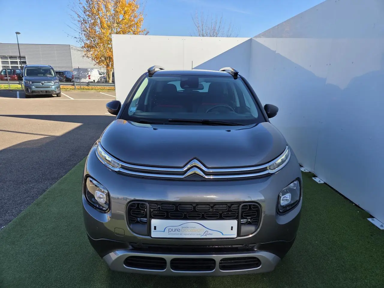 Vue de face d'un Citroën C3 Aircross gris platinium avec toit noir perlé et barres de toit argentées sur fond urbain.
