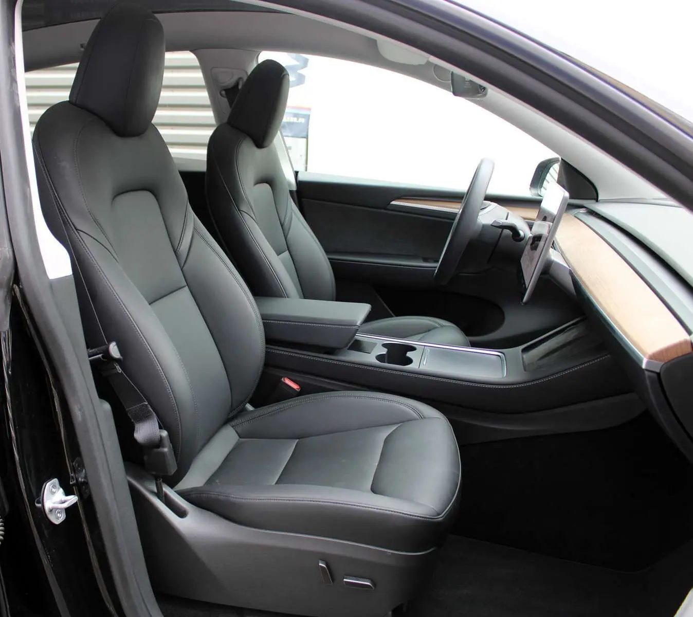 Intérieur de la Tesla Model Y 2024 noir métal, vue côté gauche sur sièges cuir noir et tableau de bord bois clair.