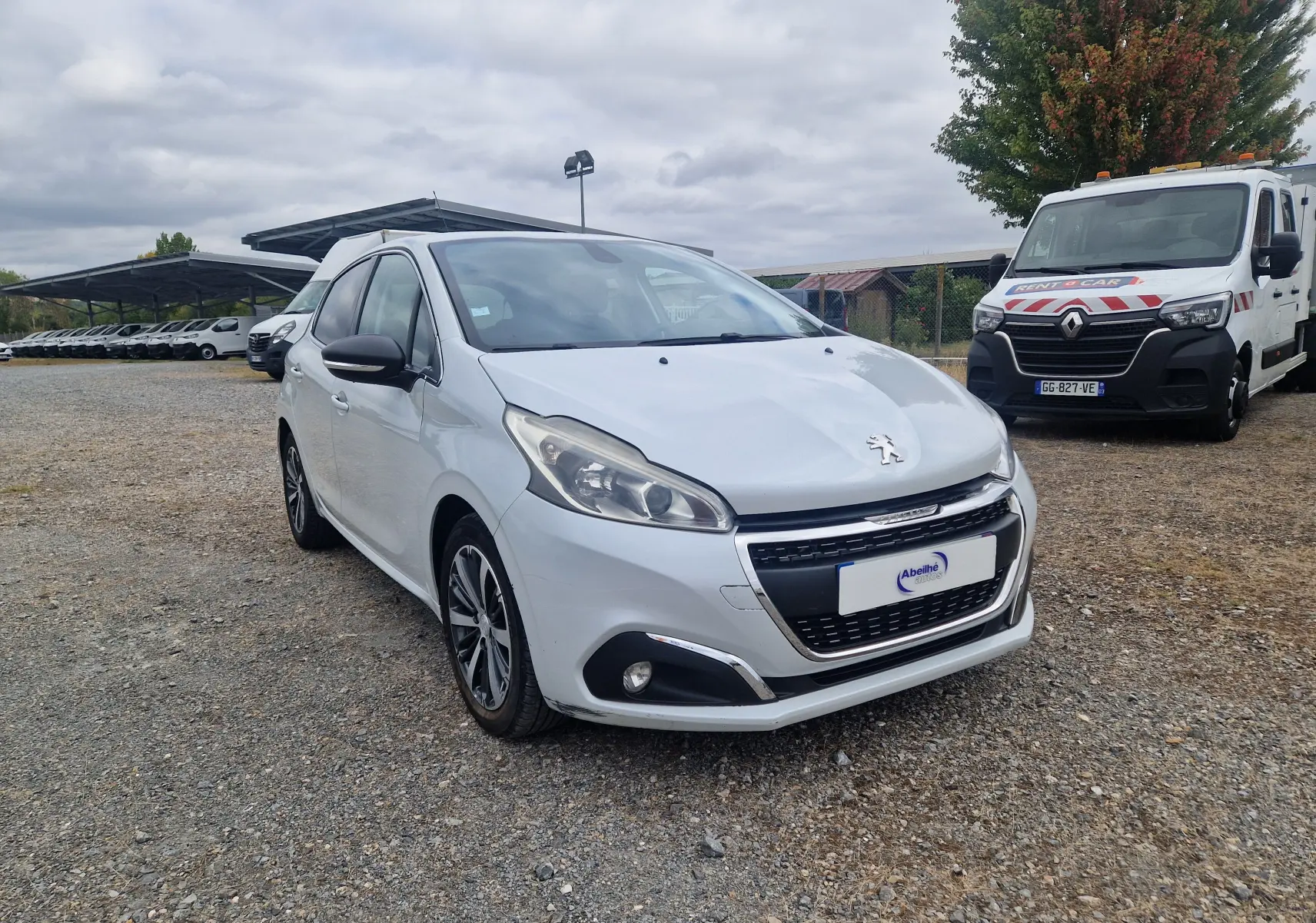 Peugeot 208 blanc nacré vue 3/4 avant droit, avec jantes alliage bicolores et calandre chromée, stationnée sur sol gravillonné.