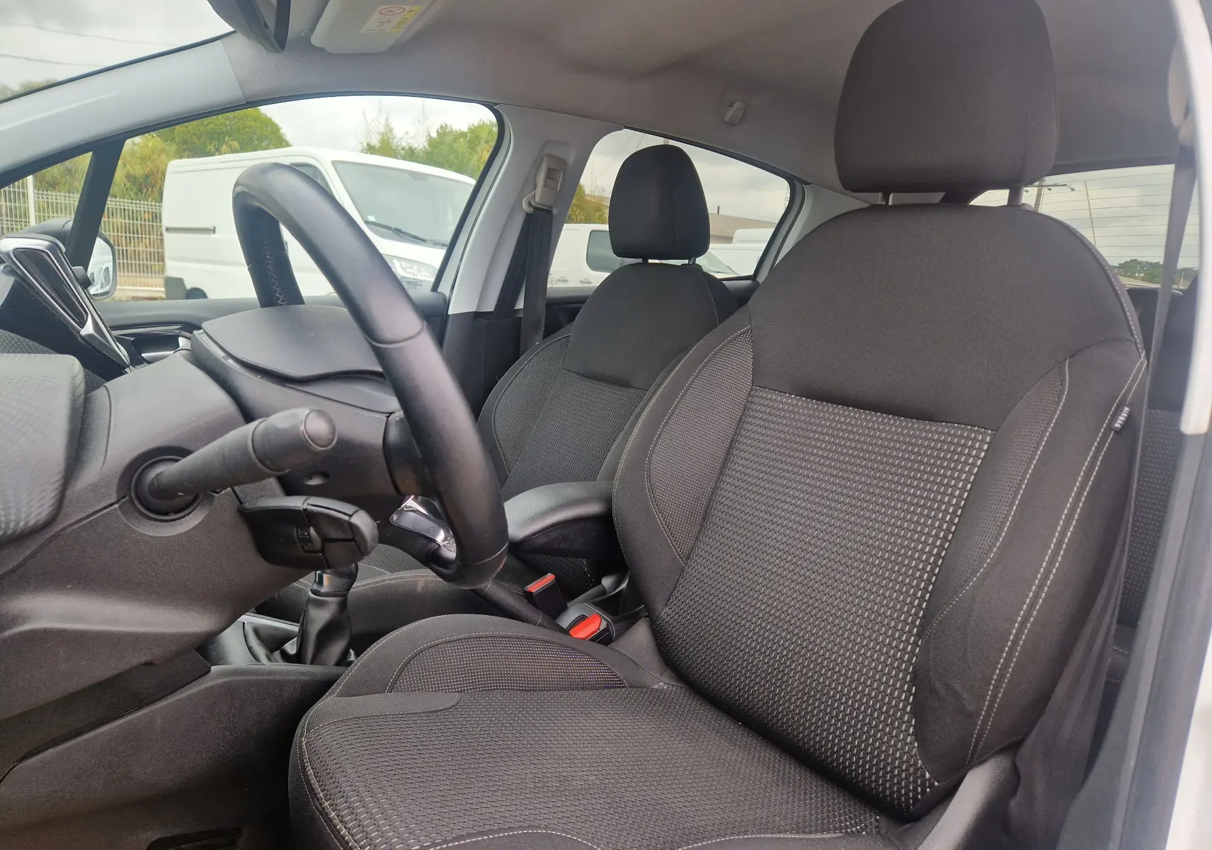 Vue intérieure côté conducteur de la Peugeot 208 blanc nacré, montrant les sièges noirs et le volant avec boîte manuelle.