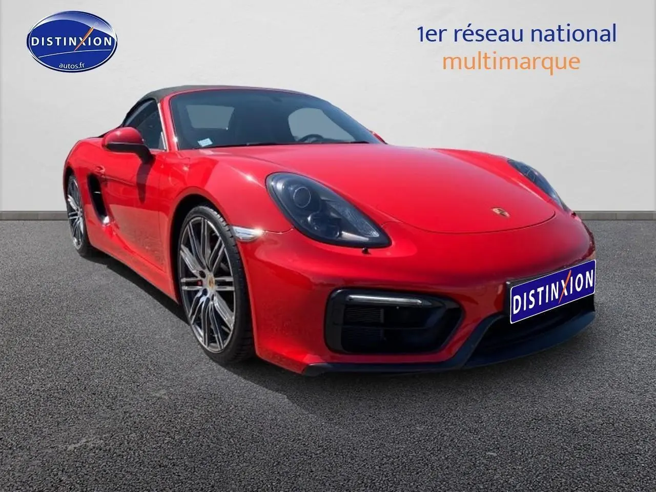 Porsche Boxster rouge vue 3/4 avant droit avec jantes alu et capote souple noire sur sol gris.