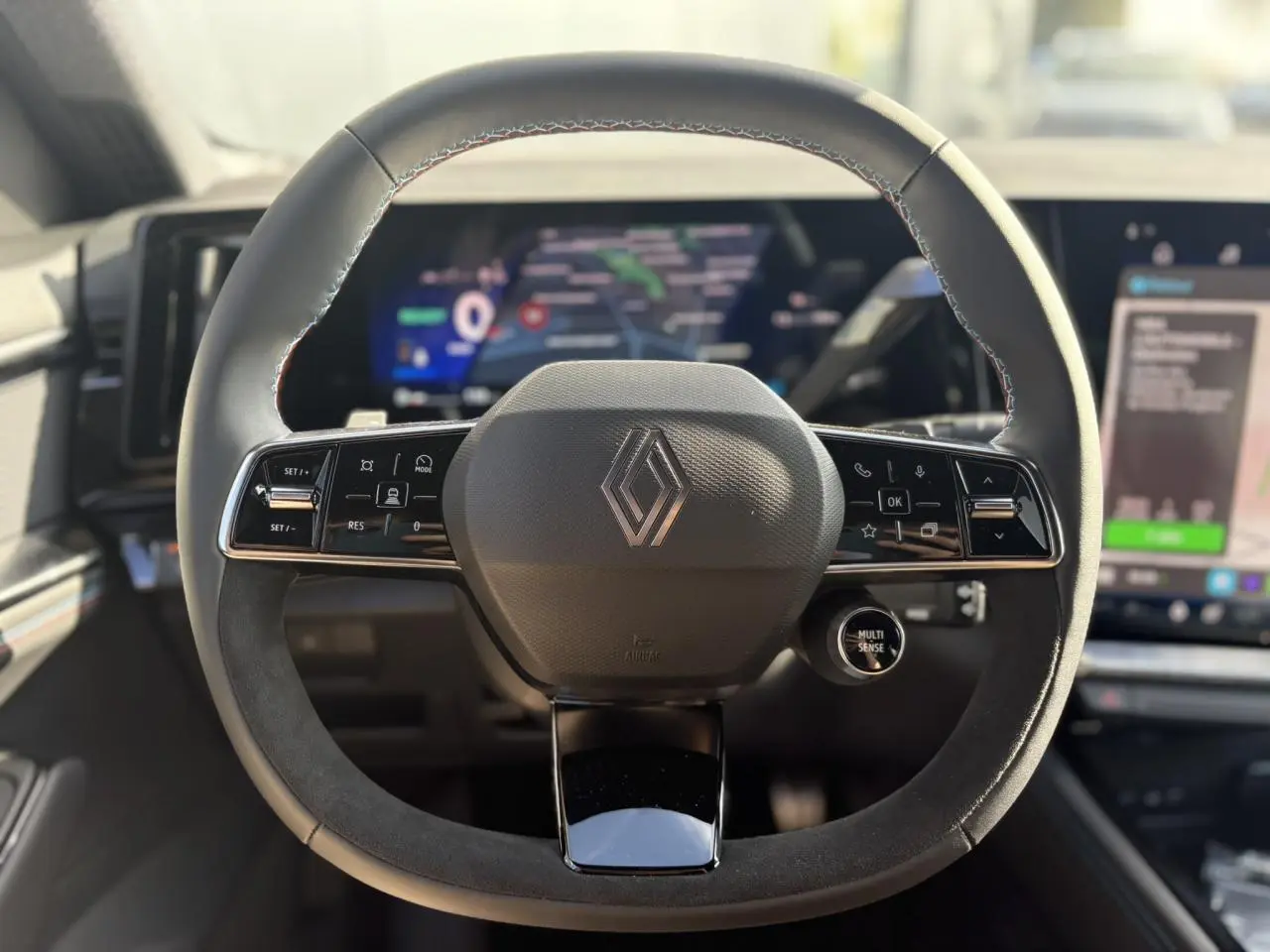 Volant Alcantara avec logo Renault et commandes intégrées, tableau de bord numérique flou en arrière-plan.