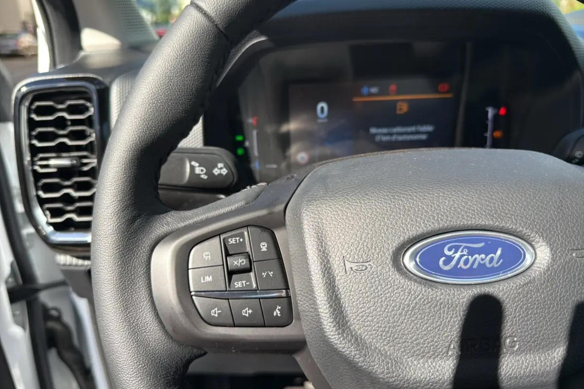 Vue rapprochée du volant cuir noir du Ford Ranger blanc glacier avec commandes multifonctions et tableau de bord numérique flou.