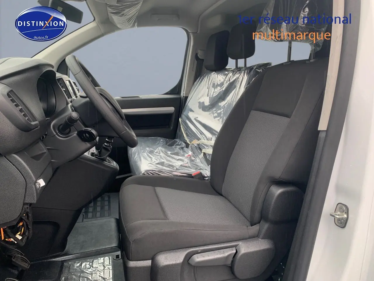 Vue intérieure côté conducteur du Peugeot Expert Long blanc Kaolin, sièges noirs avec protections plastiques visibles.