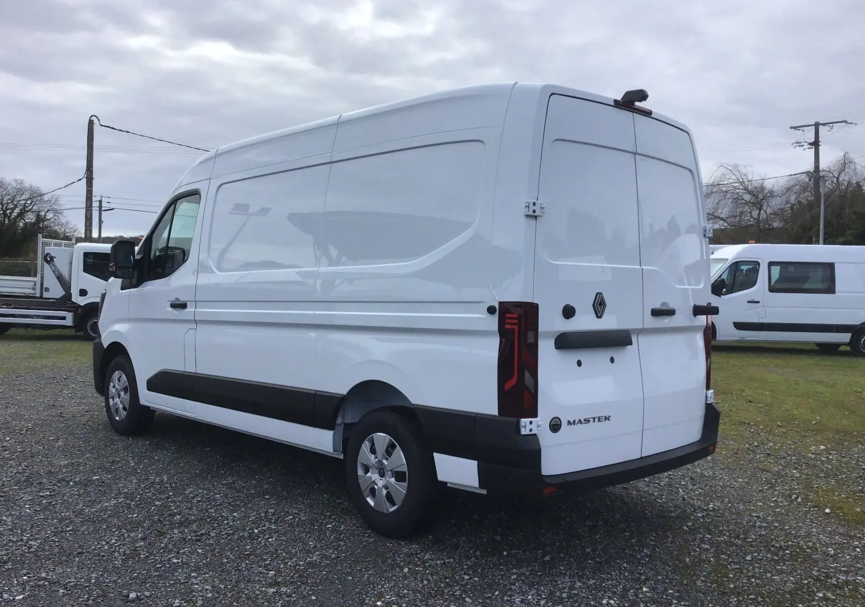 Vue 3/4 arrière droite d’un Renault Master blanc, fourgon utilitaire avec portes arrière fermées sur un sol gravillonné.