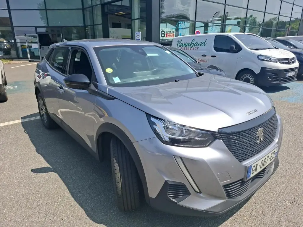 Peugeot 2008 gris Artense en 3/4 avant droit, avec calandre noire et jantes alliage foncé, stationnée sur un sol gravillonné.