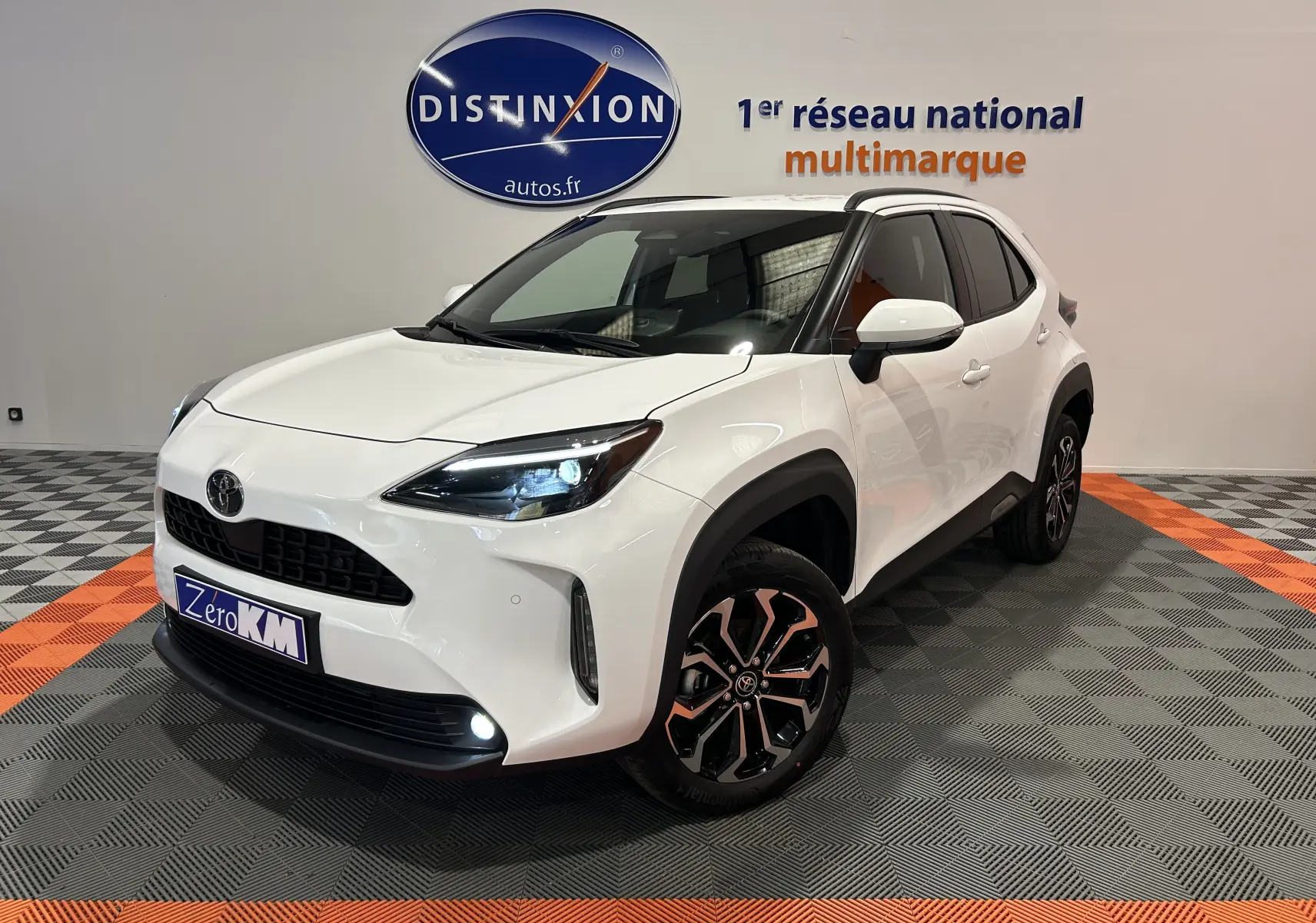 Toyota Yaris Cross blanc en 3/4 avant droit, avec jantes noires et phares LED allumés en showroom.