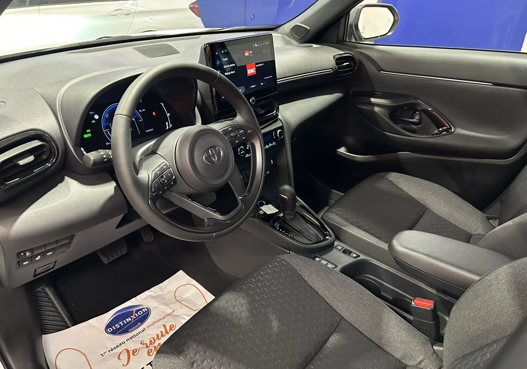 Intérieur de la Toyota Yaris Cross 130 DESIGN MY25 blanc, vue côté conducteur montrant tableau de bord et sièges tissu noirs.