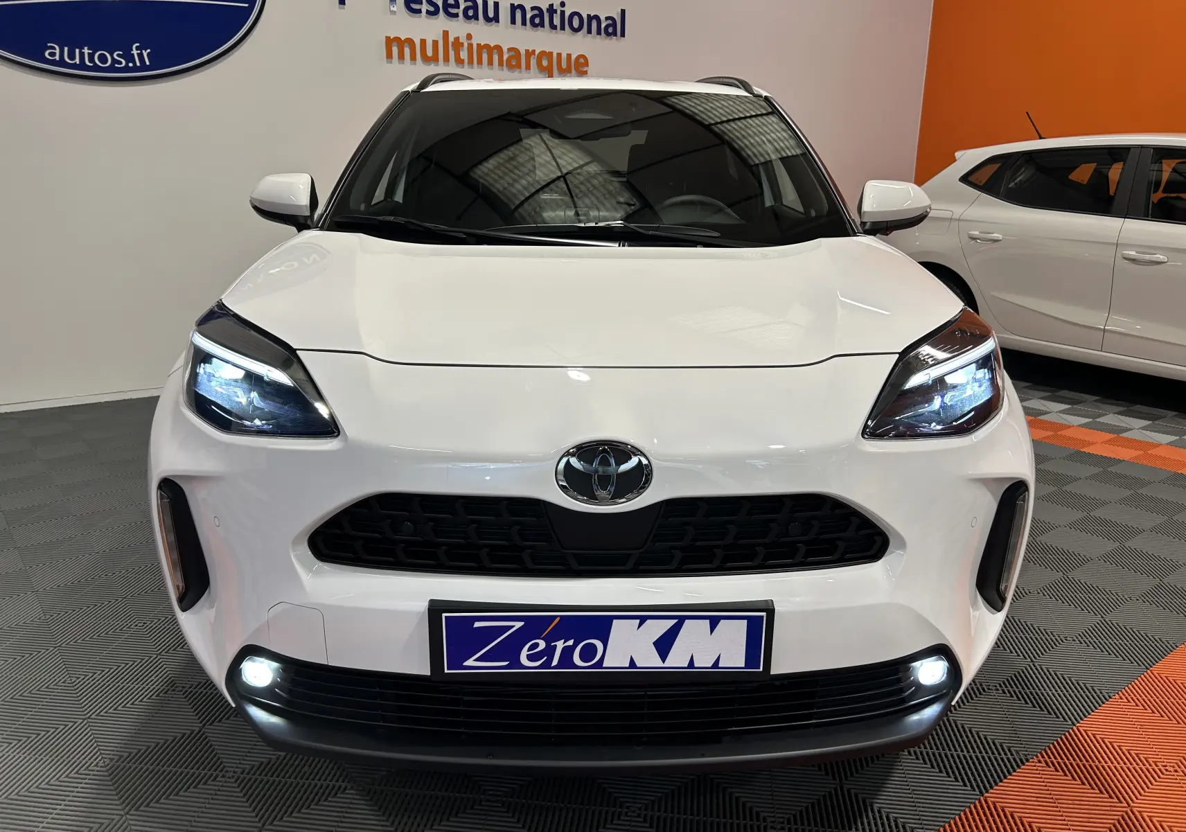 Vue avant d'une Toyota Yaris Cross blanche 2025 avec phares LED allumés et plaque Zéro KM visible.