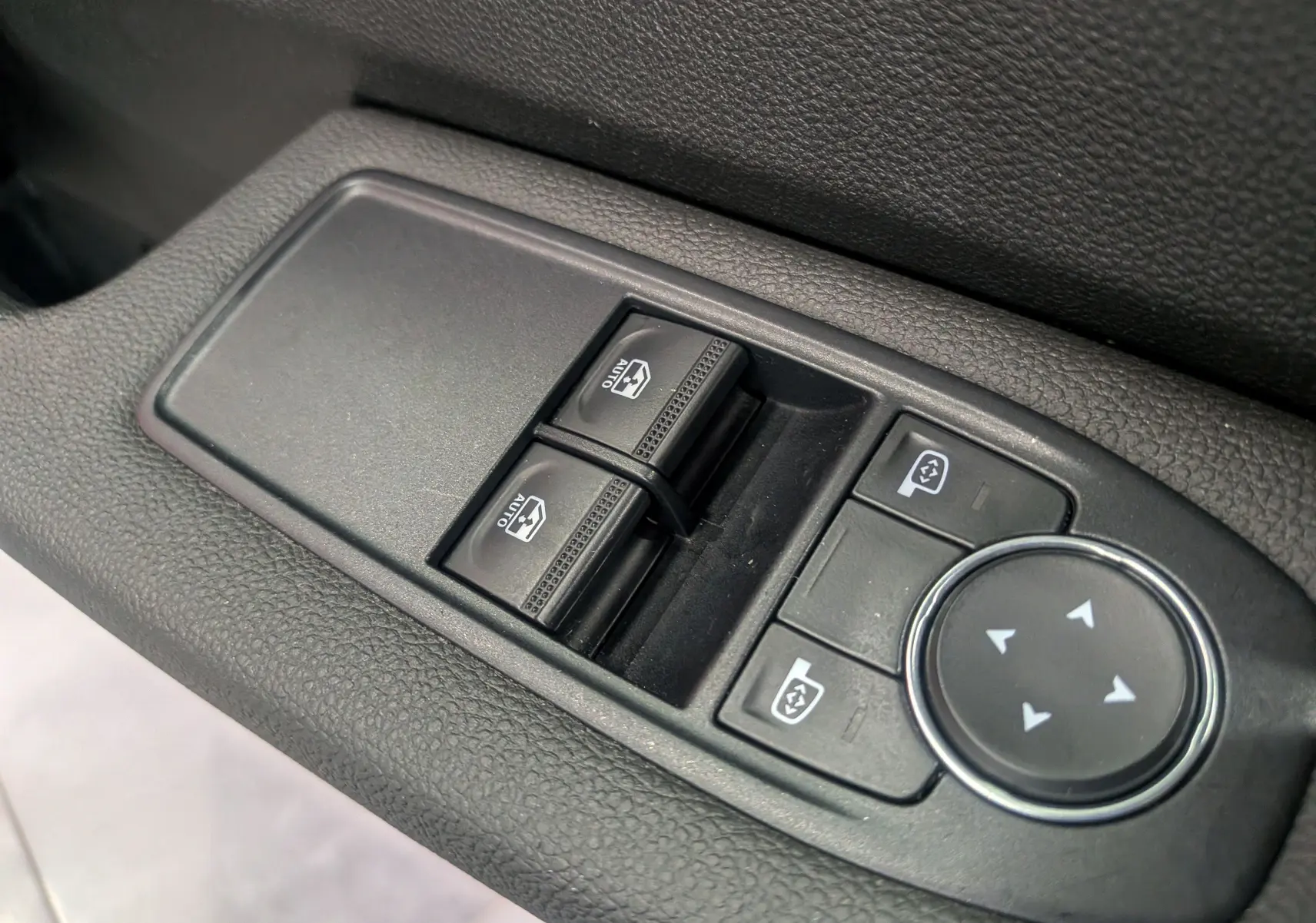 Gros plan sur les commandes des vitres électriques et réglages des rétroviseurs côté conducteur d'une Renault Clio V blanche.