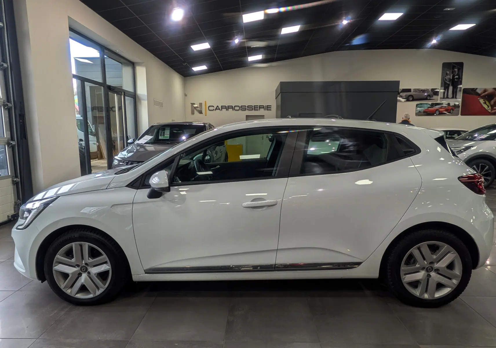 Profil latéral droit d'une Renault Clio V blanc glacier en showroom, soulignant ses jantes alliage et lignes épurées.
