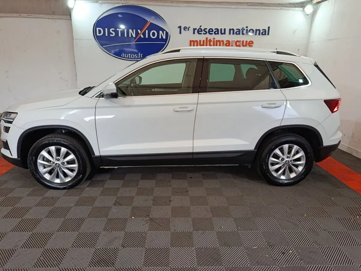 Vue de profil côté gauche d'un SKODA KAROQ blanc 2024, SUV compact avec barres de toit noires et jantes alliage.