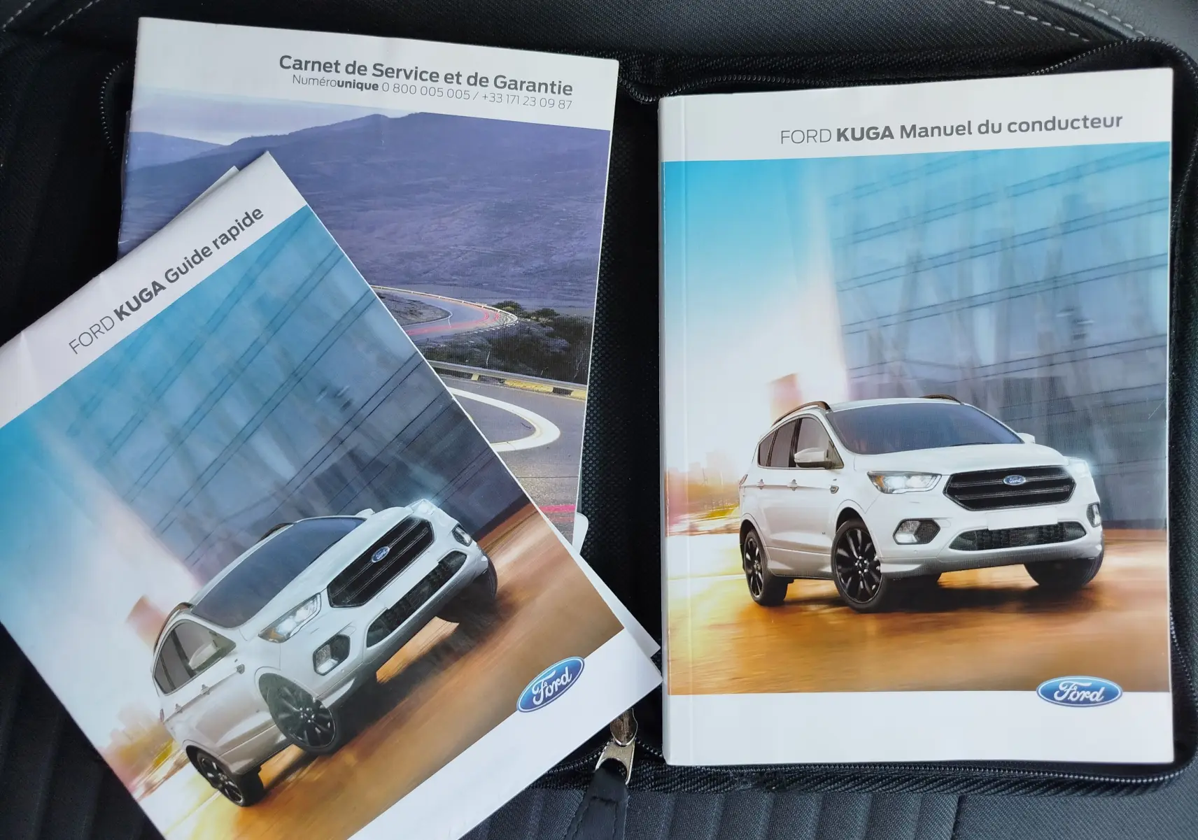 Vue 3/4 avant d'un Ford Kuga gris clair avec jantes noires sur fond urbain flouté, sur deux manuels du conducteur.