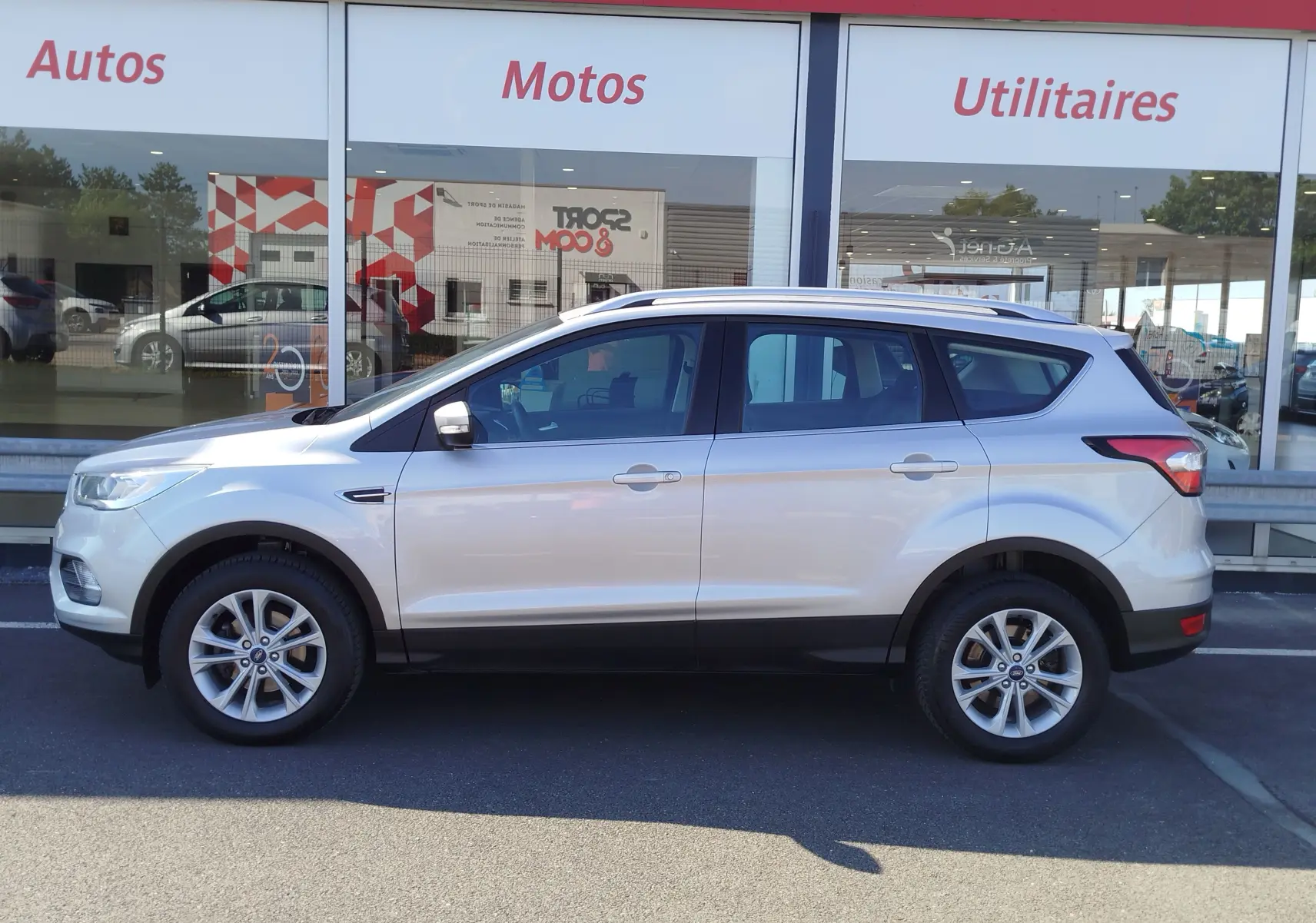 Profil côté gauche d'un Ford Kuga gris clair 2019 garé devant une vitrine de concession automobile.
