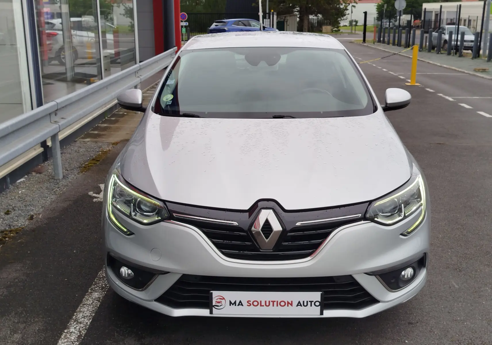 Vue avant d'une Renault Mégane gris titanium 2018 avec phares allumés et logo Renault bien visible.