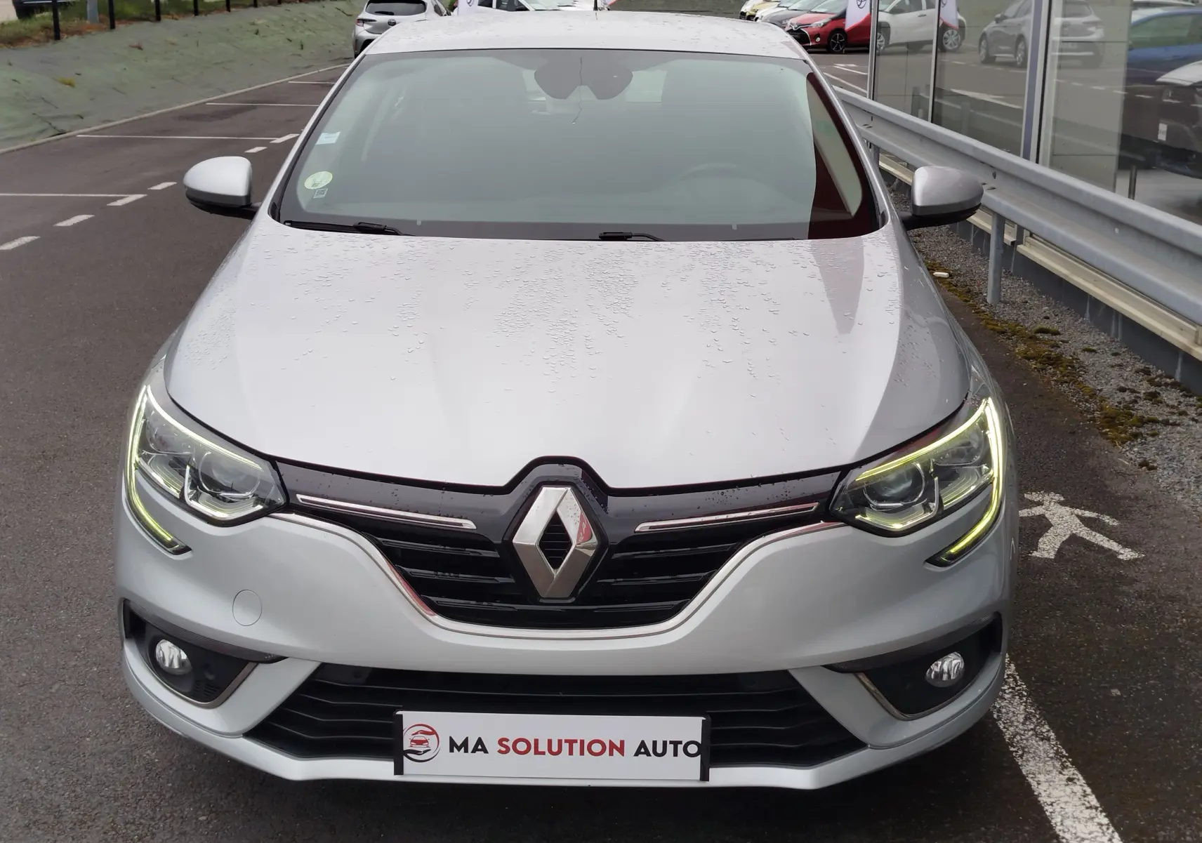 Vue frontale d'une Renault Mégane gris titanium avec phares allumés et logo Renault bien visible sur la calandre.