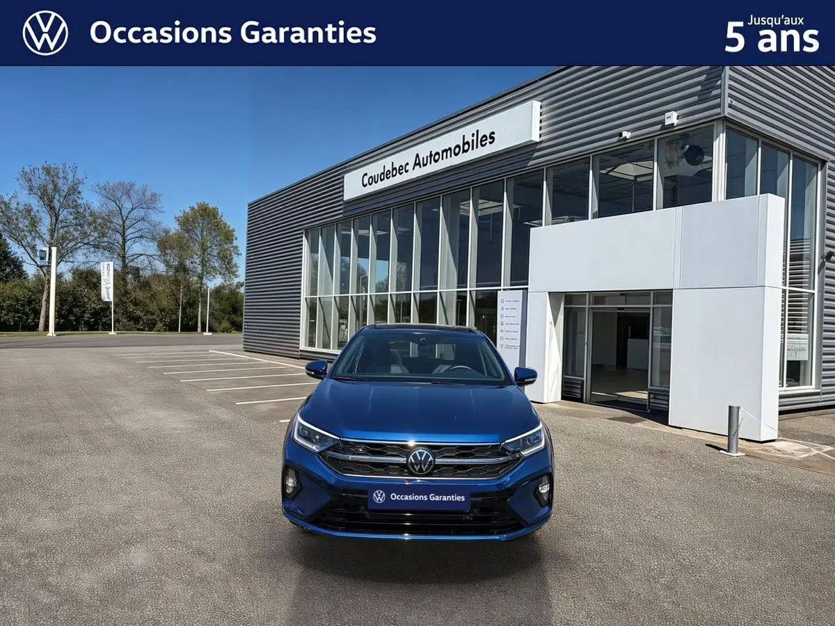 Vue frontale d'un Volkswagen Taigo bleu métallisé garé devant un bâtiment moderne de concession automobile.