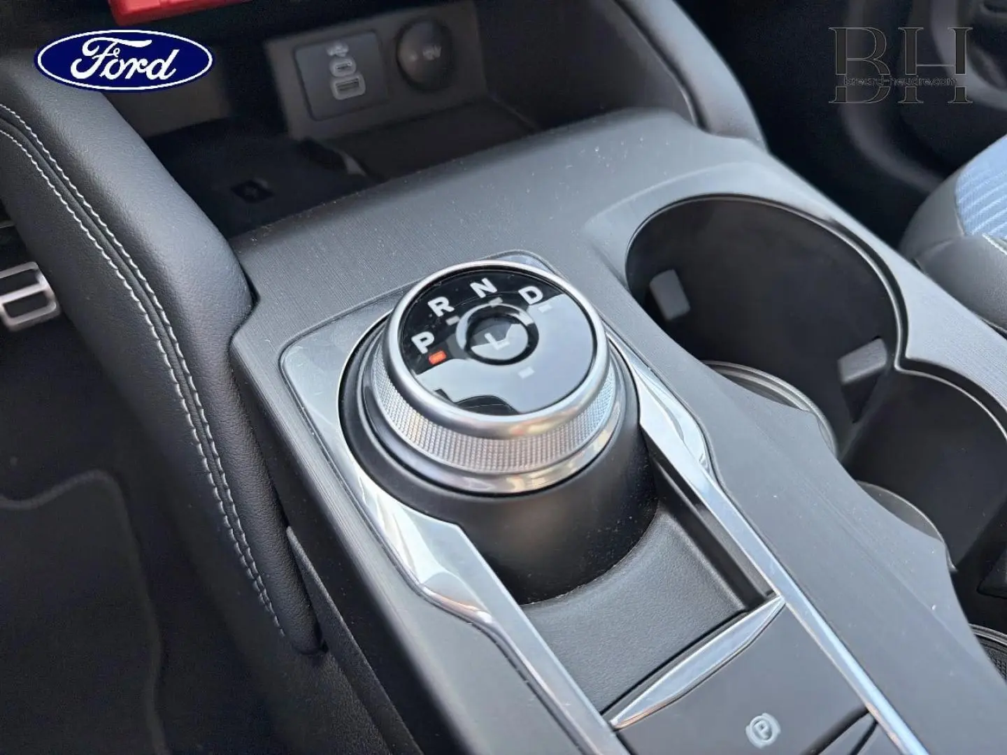 Gros plan sur la molette de boîte automatique centrale du Ford Kuga bleu avec accoudoir noir et porte-gobelets.
