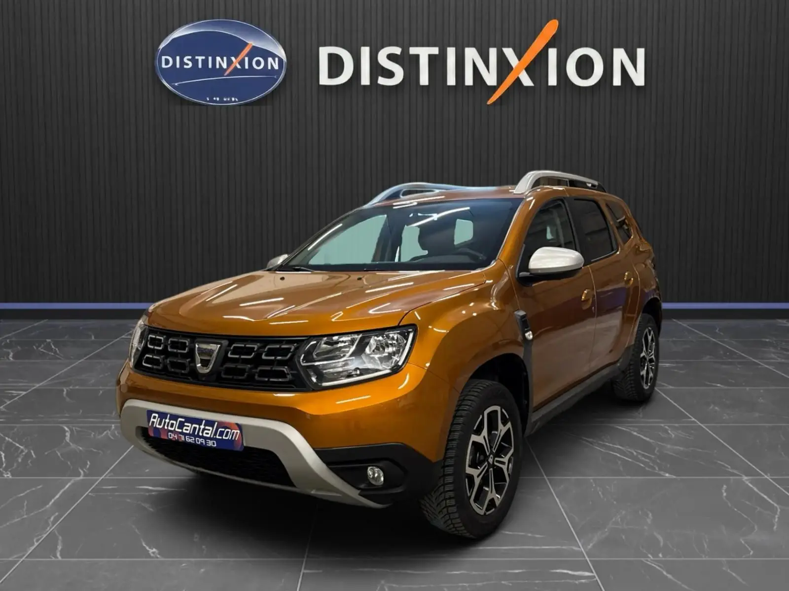 Dacia Duster orange en 3/4 avant droit, avec jantes bi-ton et rétroviseurs blancs, sur sol gris intérieur showroom.