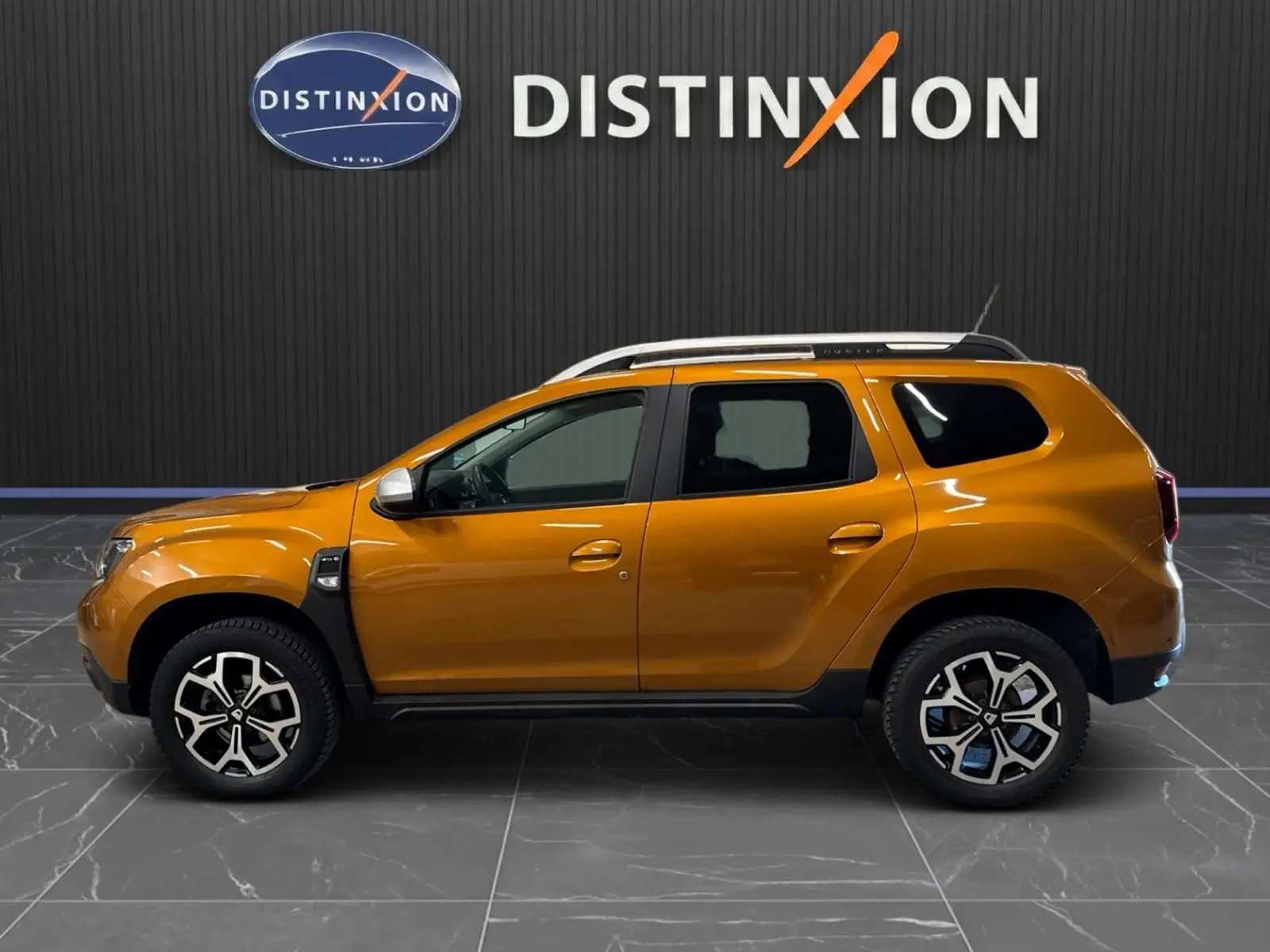 Vue de profil côté gauche d'un Dacia Duster orange 2020 avec jantes bi-ton et barres de toit blanches.