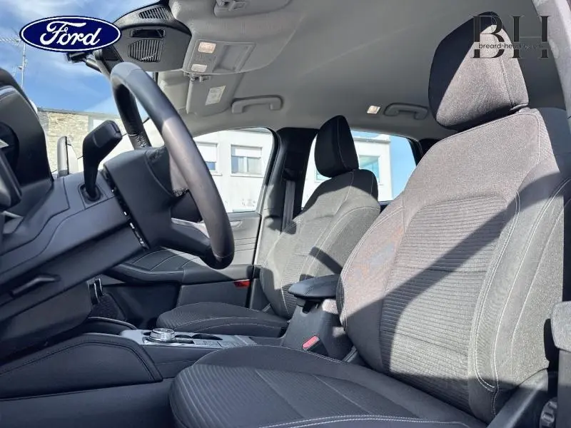 Vue intérieure côté conducteur du Ford Kuga 2023, sièges sport en tissu gris avec accoudoir central et volant cuir multifonction.