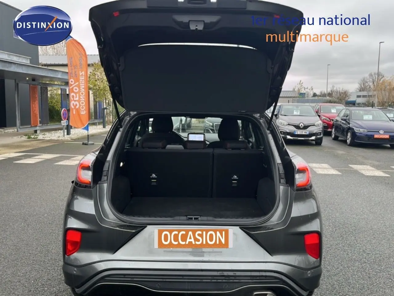 Vue arrière d'un Ford Puma gris Magnetic Metal 2024 avec coffre ouvert sur un parking de voitures d'occasion.