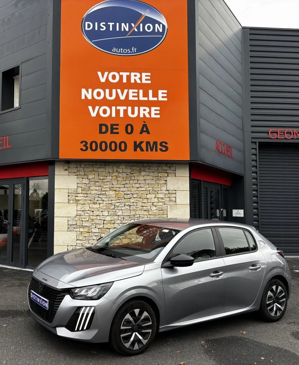 Peugeot 208 PureTech 100 S&S Active gris clair vue 3/4 avant droit devant un bâtiment avec panneau orange Distinxion.
