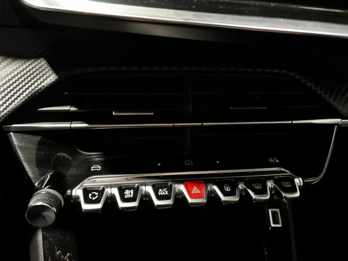 Gros plan sur la rangée de boutons métalliques du tableau de bord du Peugeot 208 gris clair 2024, avec bouton rouge d'alerte au centre.