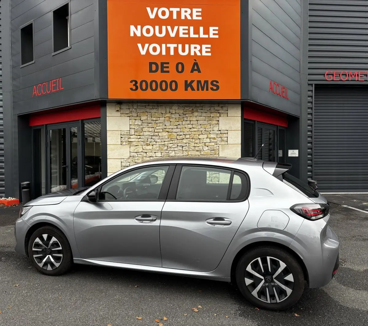 Peugeot 208 PureTech 100 S&S Active gris clair vue de profil côté gauche devant un bâtiment avec panneau orange "Votre nouvelle voiture".