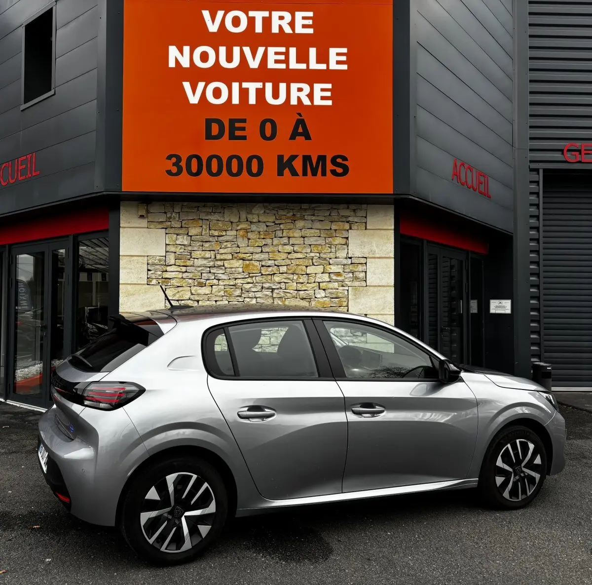 Peugeot 208 gris clair vue de profil côté droit devant un mur en pierre avec panneau orange publicitaire.