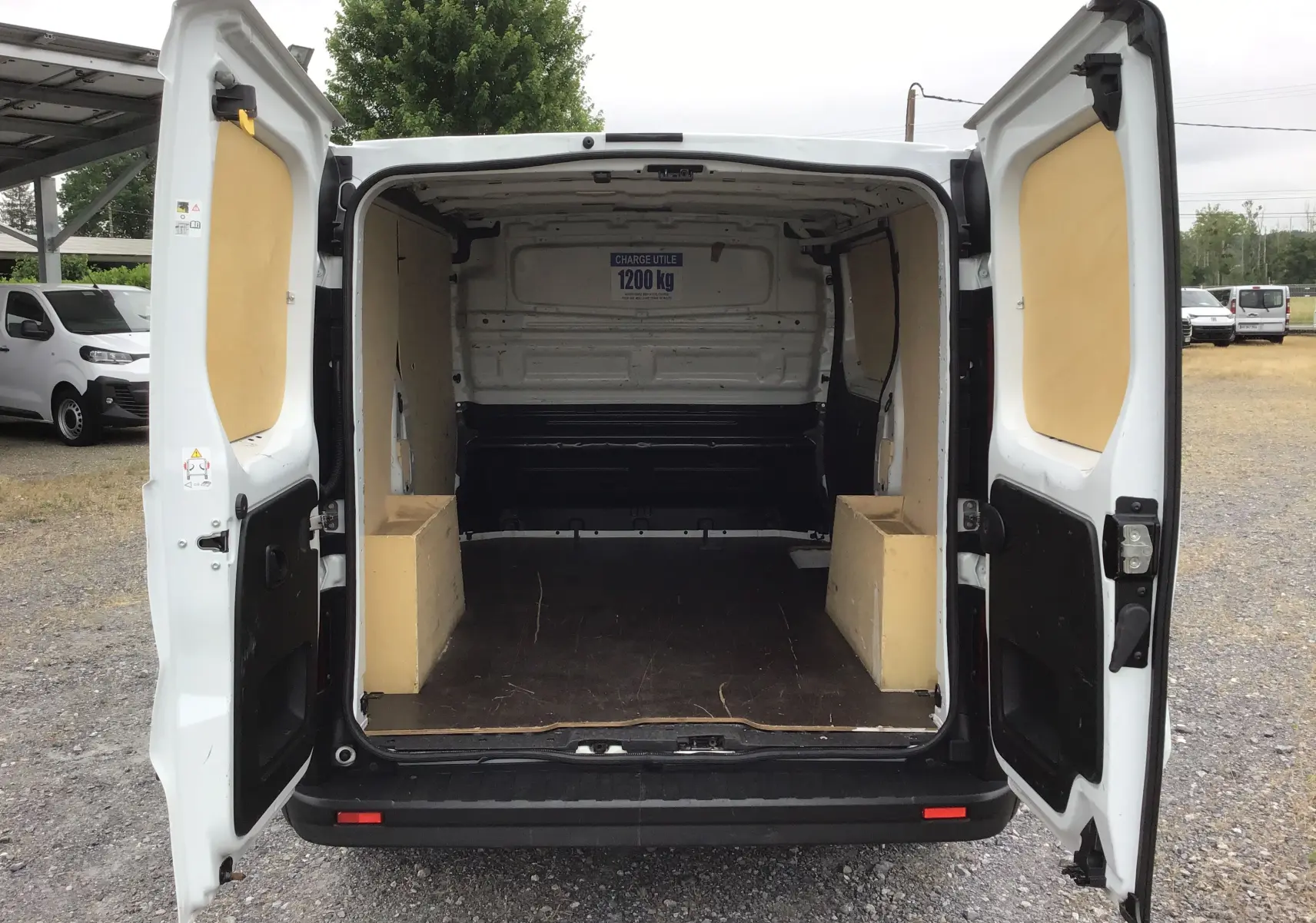 Vue arrière du Renault Trafic Fourgon blanc 2024 avec portes ouvertes montrant l’espace de chargement vide et protégé par des panneaux bois.