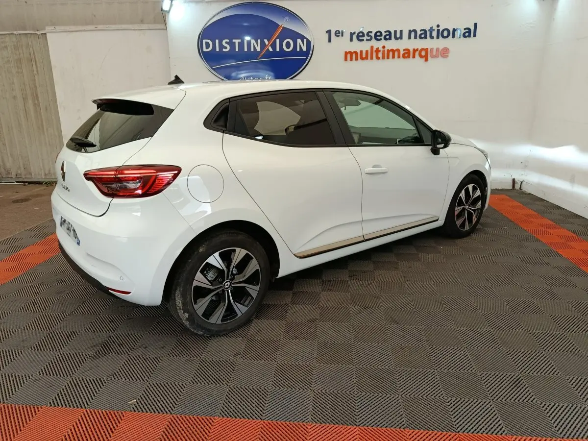 Renault Clio blanc vue 3/4 arrière droit, avec jantes noires et toit avec spoiler, en intérieur showroom.