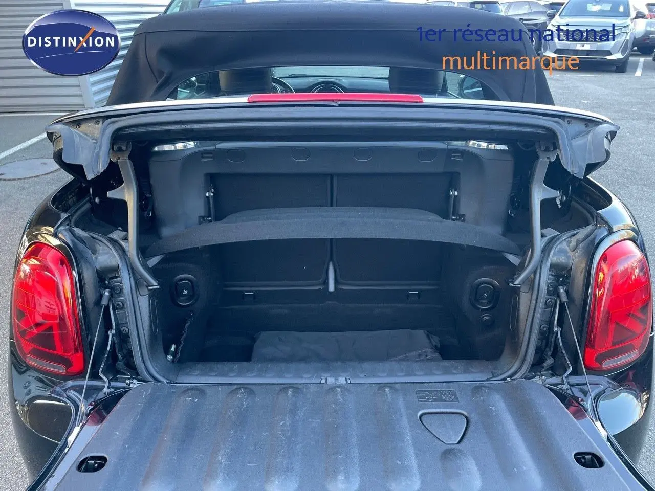 Coffre ouvert vu de l'arrière d'une MINI Cabriolet F57 noire Midnight Metal avec capote relevée sur parking.