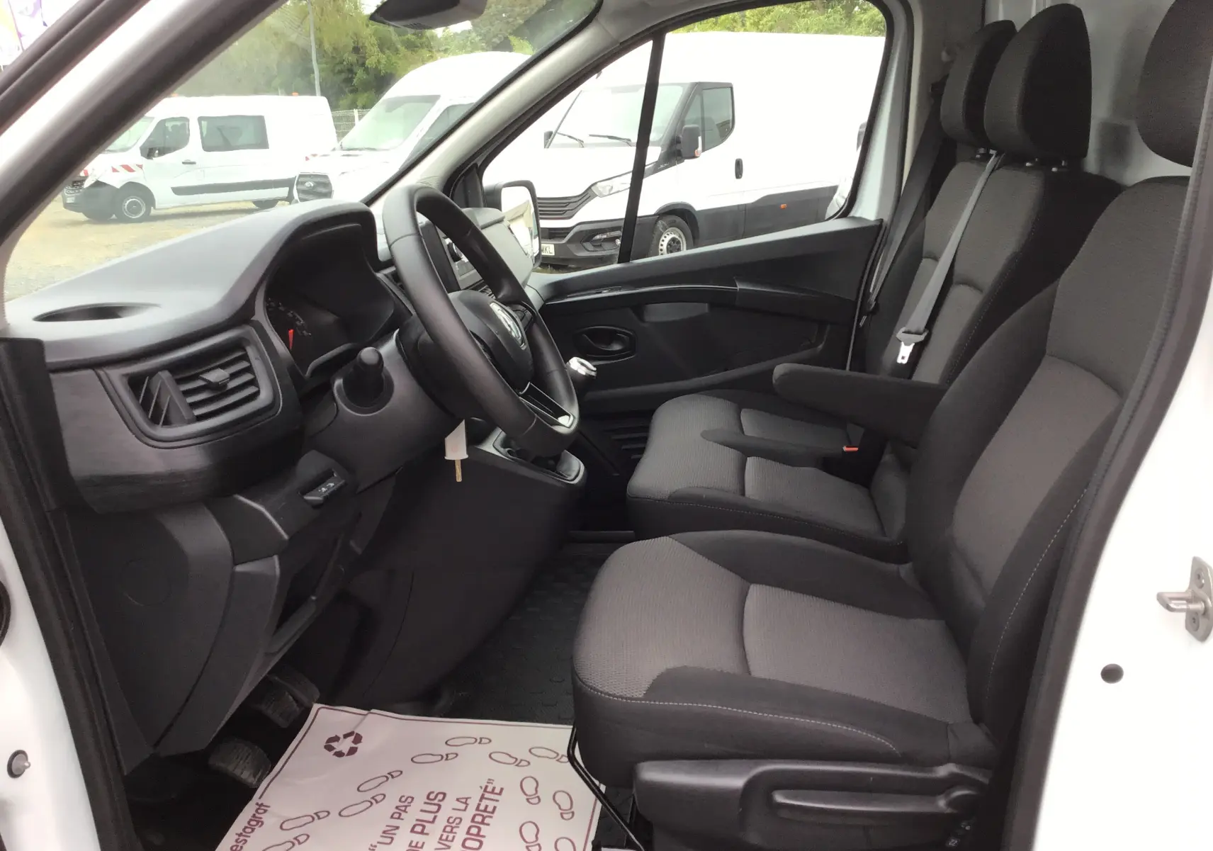 Vue intérieure côté conducteur du Renault Trafic Fourgon blanc 2024, montrant les sièges avant et le tableau de bord avec volant multifonction.